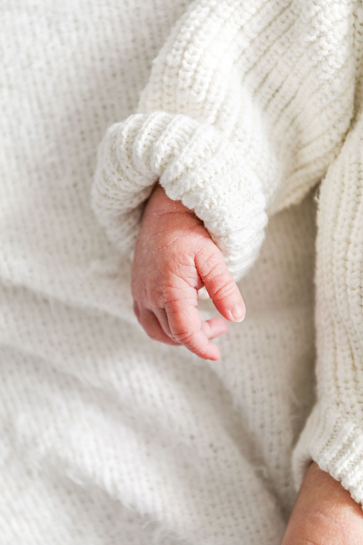 A tiny baby's hand emerges from a cozy white knit sweater sleeve against a soft white textile background, during newborn shoot in Nottingham