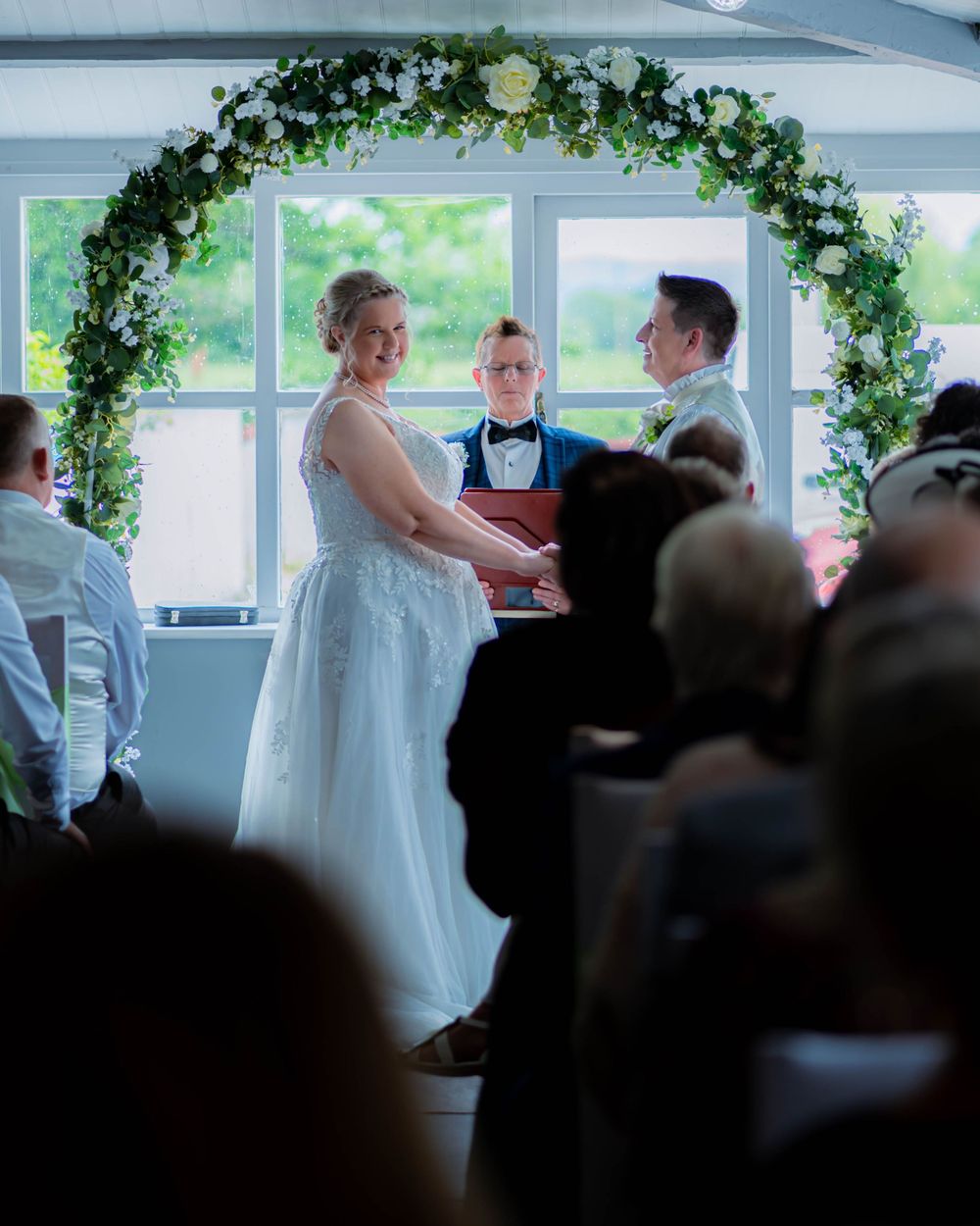 A wedding ceremony in a bright indoor venue with a beautiful floral arch and gathered guests seated in rows.