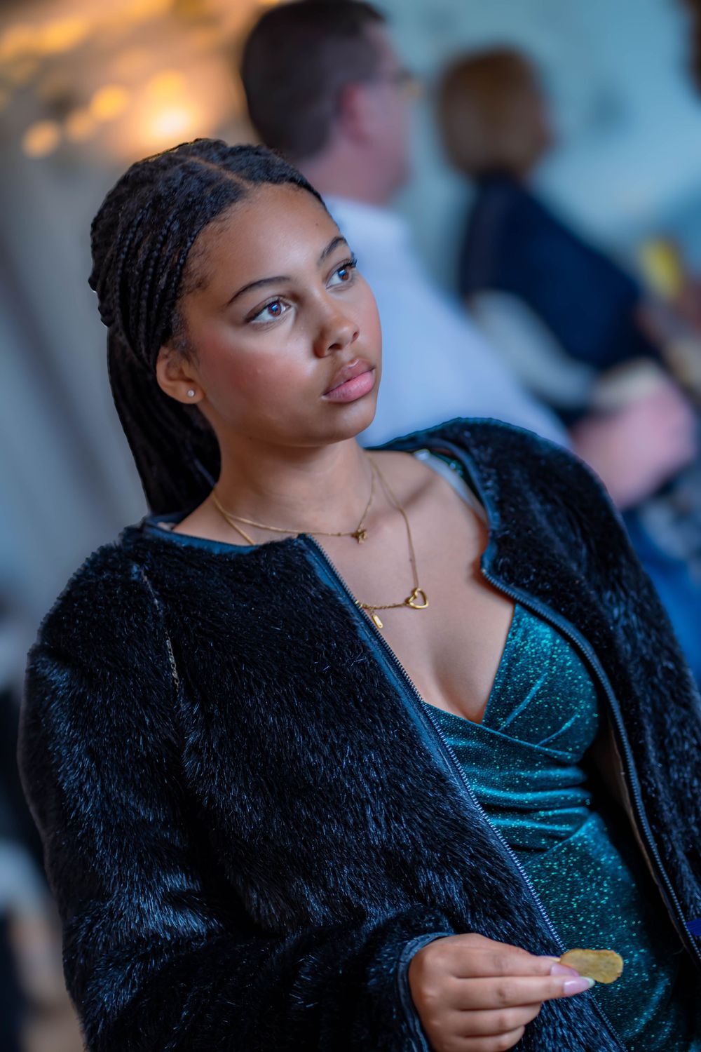 A woman in a teal velvet dress and black fur coat sits thoughtfully at an evening event.