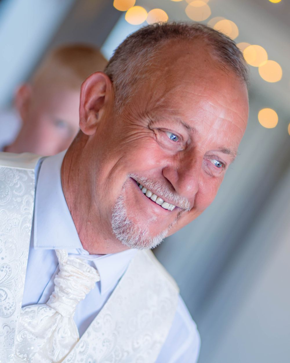Smiling person in formal white clothing with a decorative tie poses against a blurred background with bokeh lights.