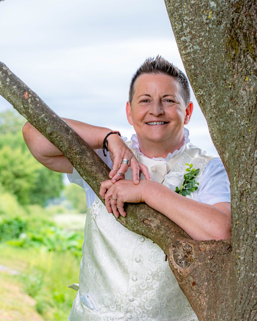 A person in white wedding attire leans against a tree branch in an outdoor garden setting.