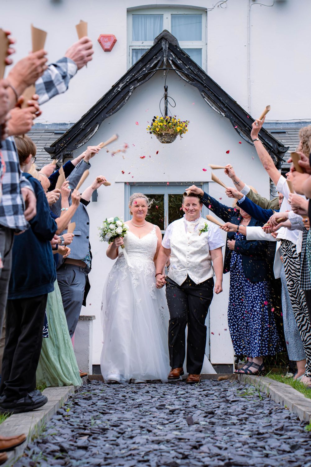 Wedding celebration with couple walking through archway while guests throw confetti along cobblestone pathway.