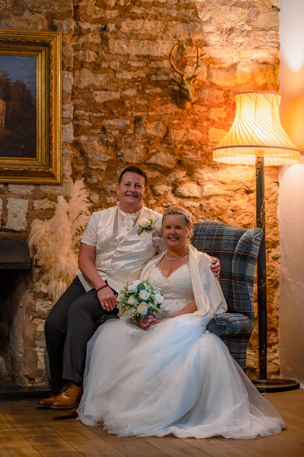 A newlywed couple relaxes on a vintage couch against an exposed brick wall with warm lighting and floral bouquet.