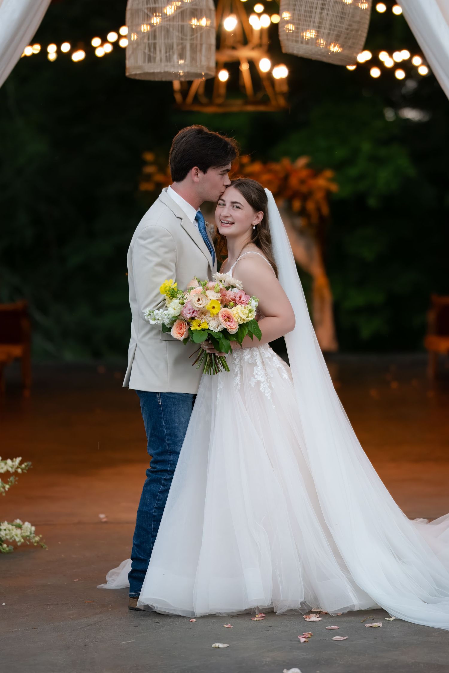 A happy newlywed couple shares an intimate moment under a canopy decorated with string lights while holding a colorful bouquet.