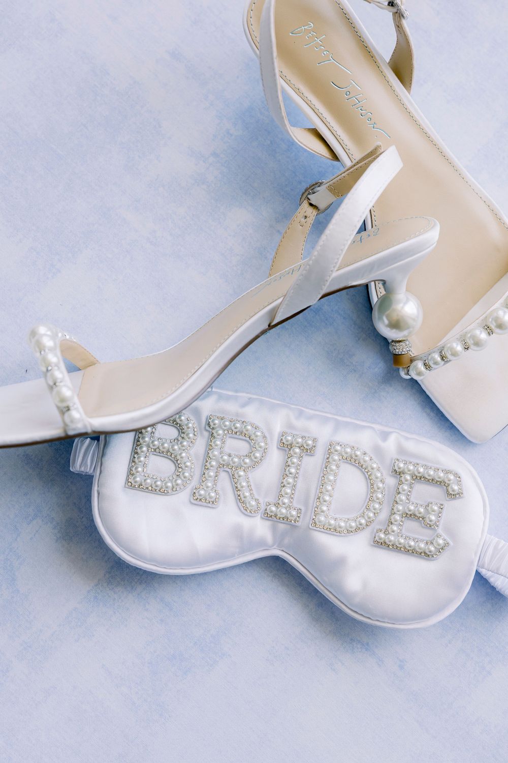 Elegant white bridal heels with pearl details next to a sparkly white sleep mask with 'BRIDE' text in rhinestones.