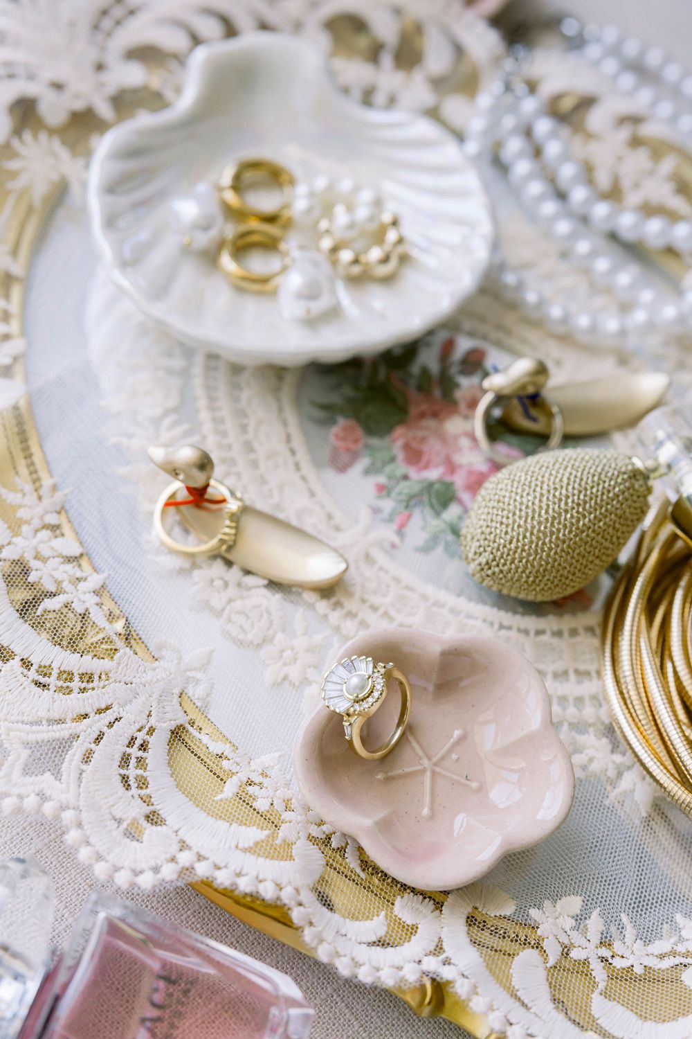 Elegant bridal accessories including gold rings, pearl necklace and perfume bottle arranged on delicate lace fabric.