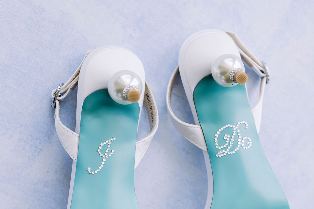 Elegant white bridal shoes with pearl details and monogrammed turquoise insoles on light blue background.