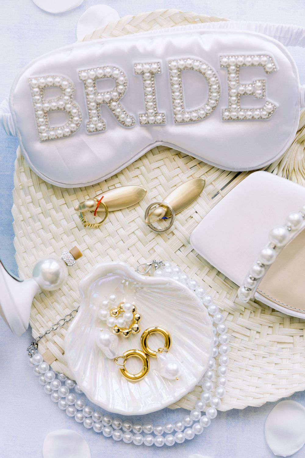 Bridal accessories including pearl-adorned sleep mask, jewelry, and rings arranged on a textured white surface.