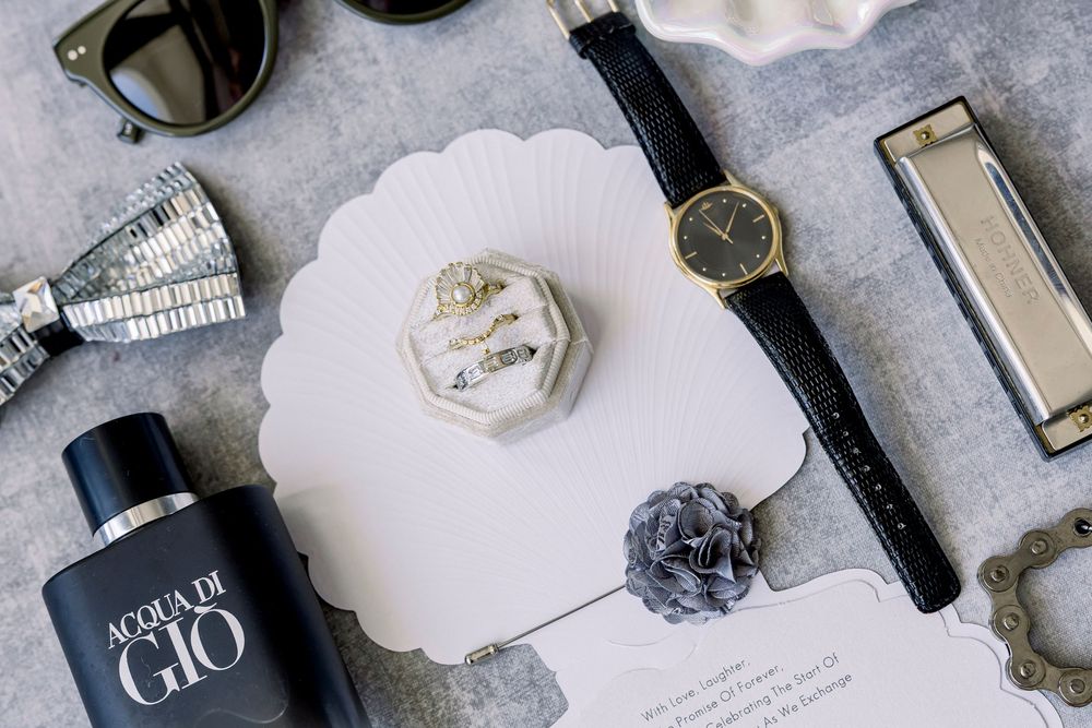 Wedding details flatlay with perfume bottle, watch, and ring box on gray surface.