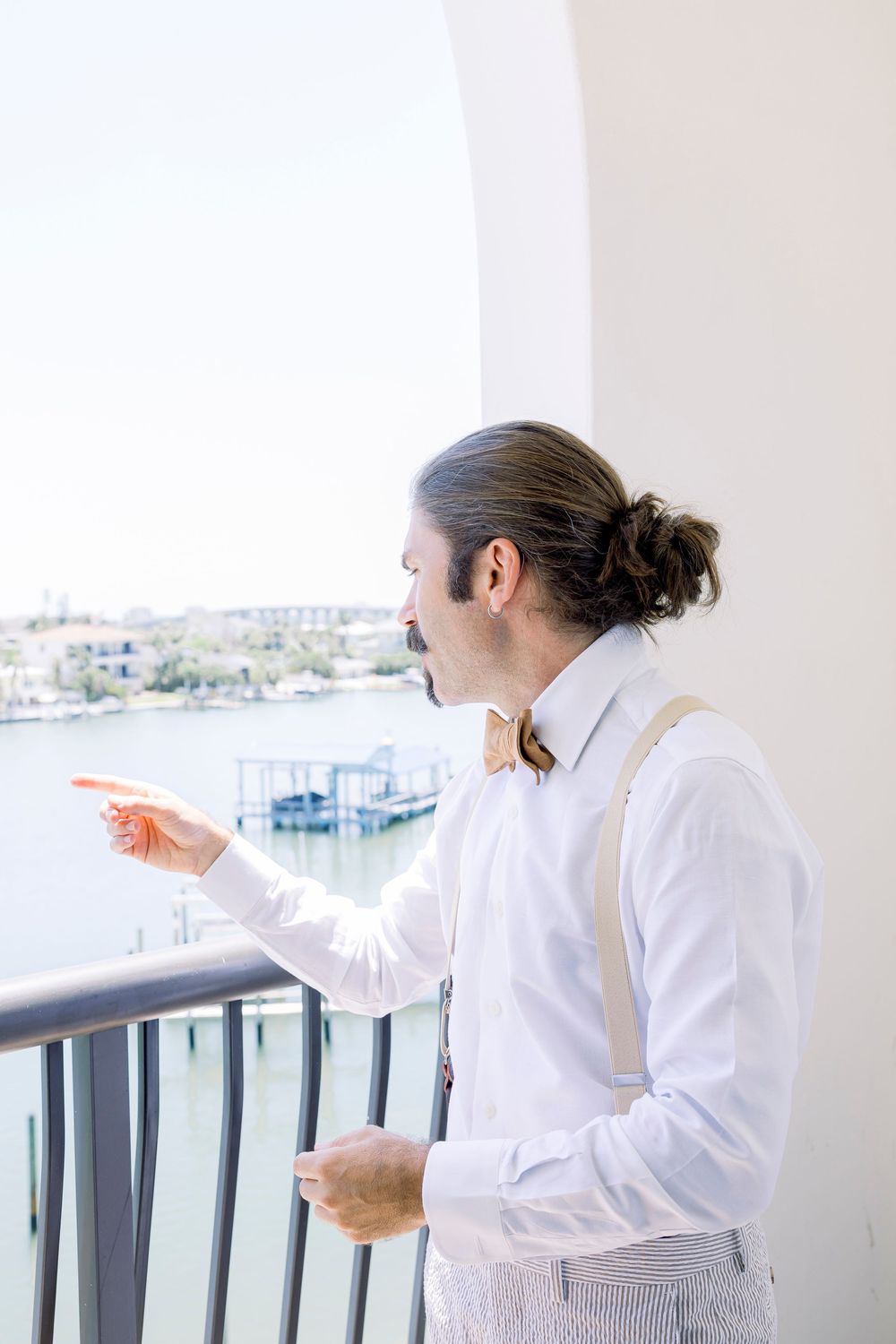 Figure in white shirt and suspenders pointing while standing on balcony overlooking marina.