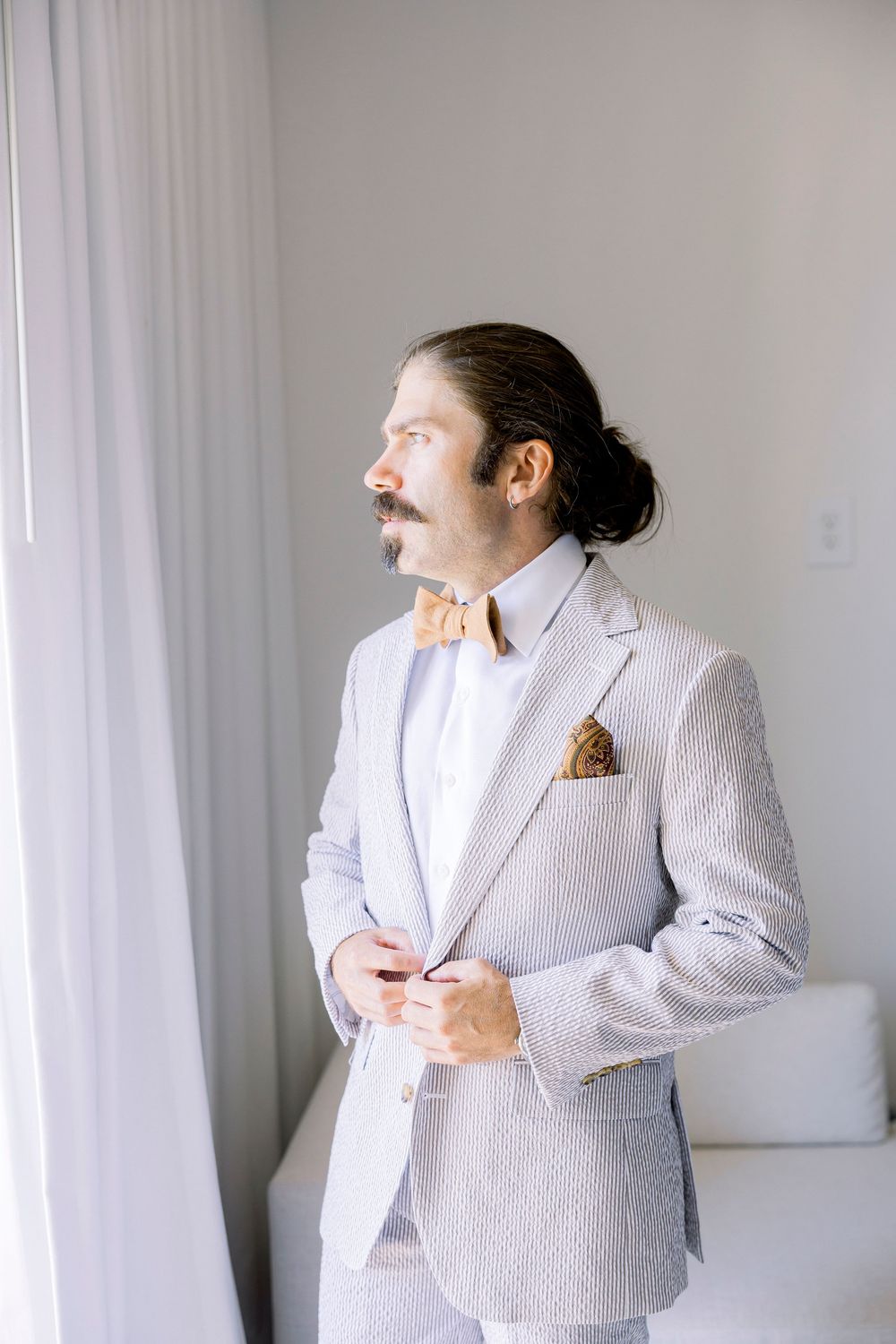 A person in a light grey textured suit with bow tie and facial hair looking to the side by a window.