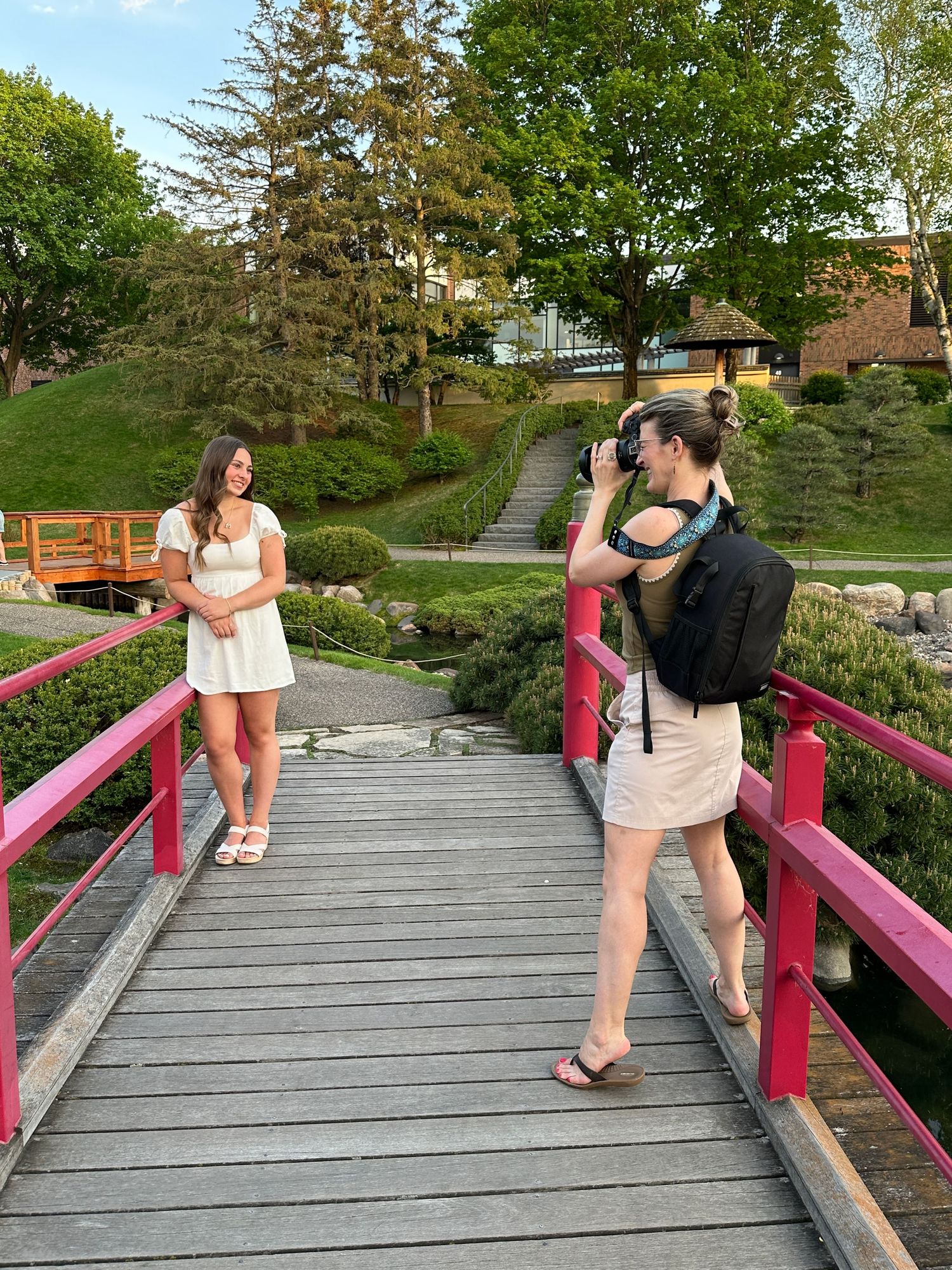 A photo session takes place on a red-railed wooden bridge in a scenic garden setting with trees and landscaping.