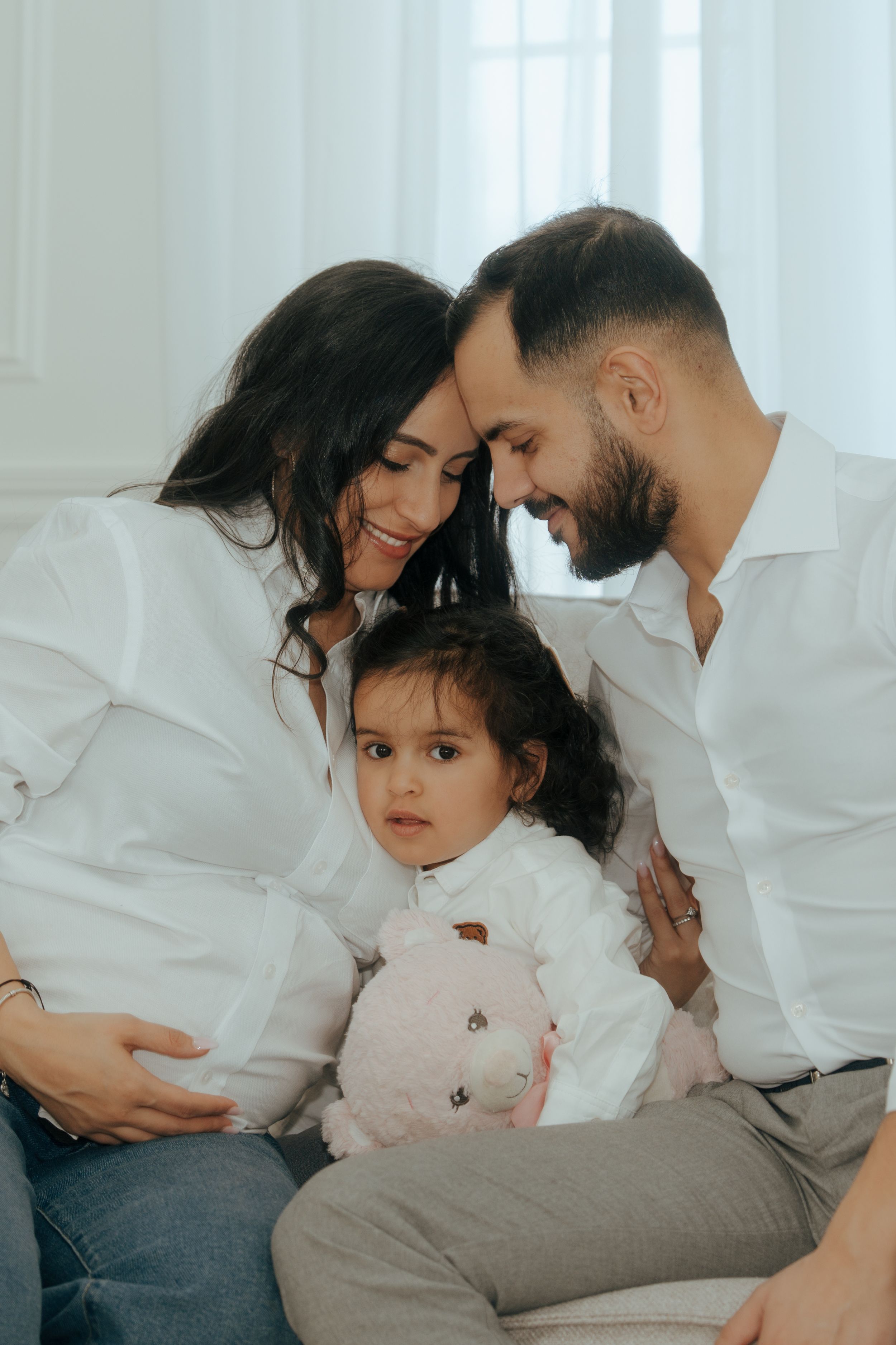 A loving family shares an intimate moment together in white clothing while sitting on a couch with their small child.