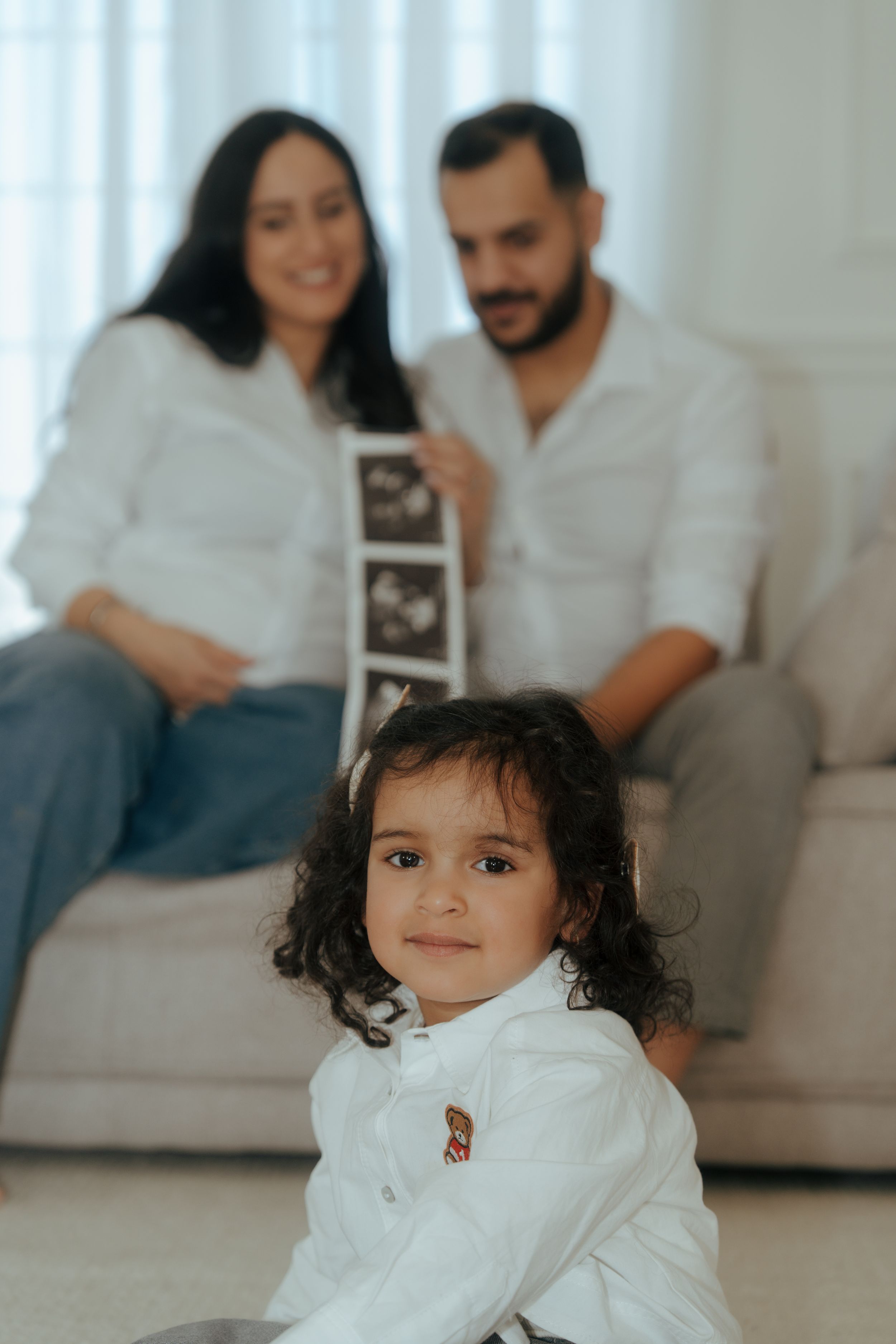 Family looks at ultrasound photos while toddler in white shirt sits on floor in living room.