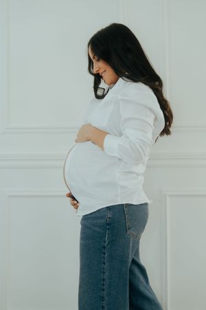 Side view of an expectant mother in jeans and white sweater lovingly holding her baby bump.