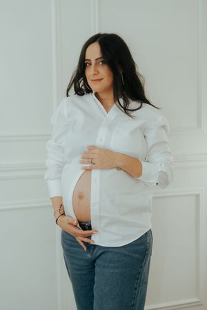 Expectant mother in white button-down shirt and jeans cradles her baby bump against white wall.