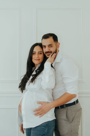 A couple dressed in white poses together against a white wall while expecting a baby.