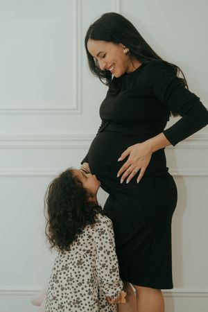 A pregnant woman in a black dress shares a tender moment with her young child against a white wall.