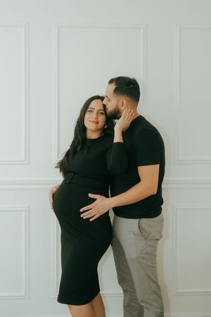 A pregnant woman in a black dress embraces with her partner against a minimalist white background.
