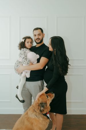 A family portrait with a small child and golden retriever dog against a white wall background.