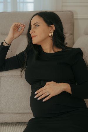 A pregnant woman in a black dress relaxes on a beige couch while touching her hair and looking to the side.
