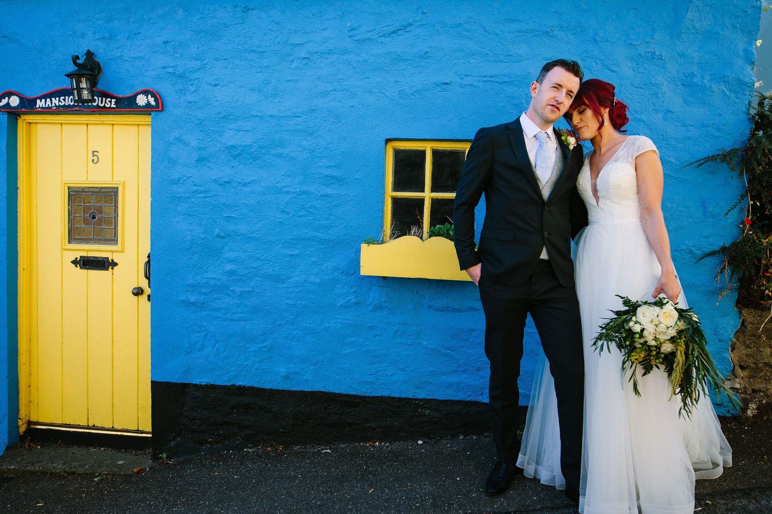 A couple elopes to Cork in Ireland poses against a vibrant blue wall with a yellow door and window frame during their wedding celebration.