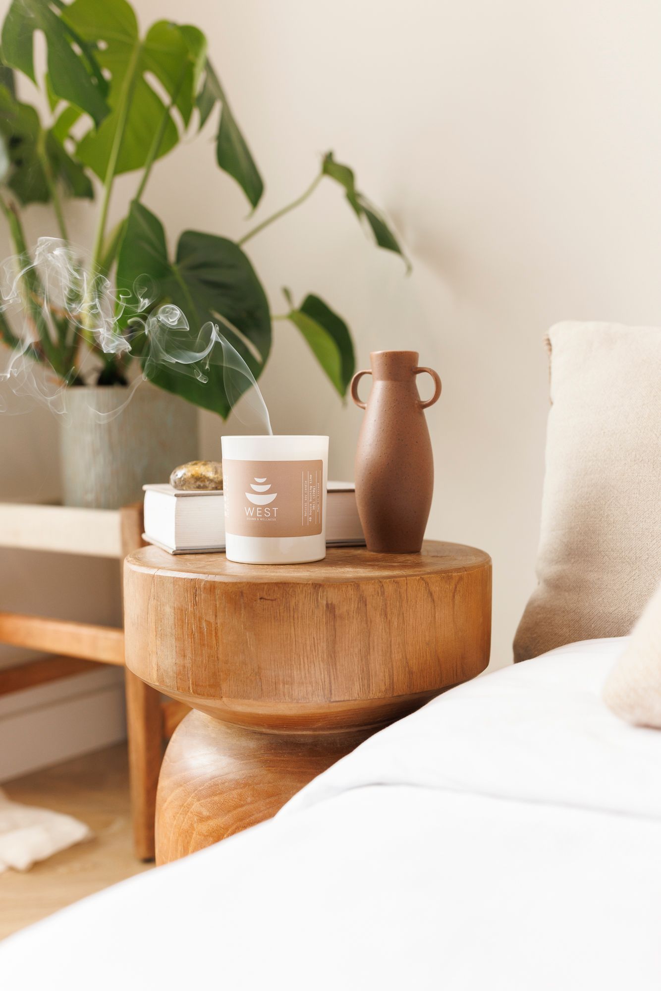 Cozy minimalist bedroom corner with wooden side table, brown ceramic vase, and monstera plant against white wall.