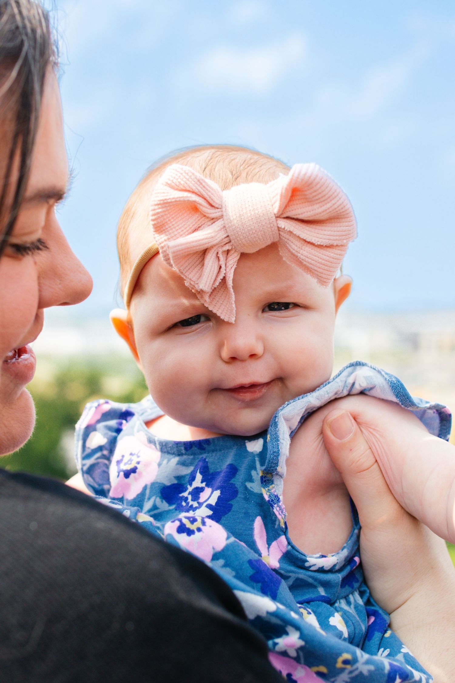 Close-up of baby wearing pink bow and floral outfit smiling against blue sky background.