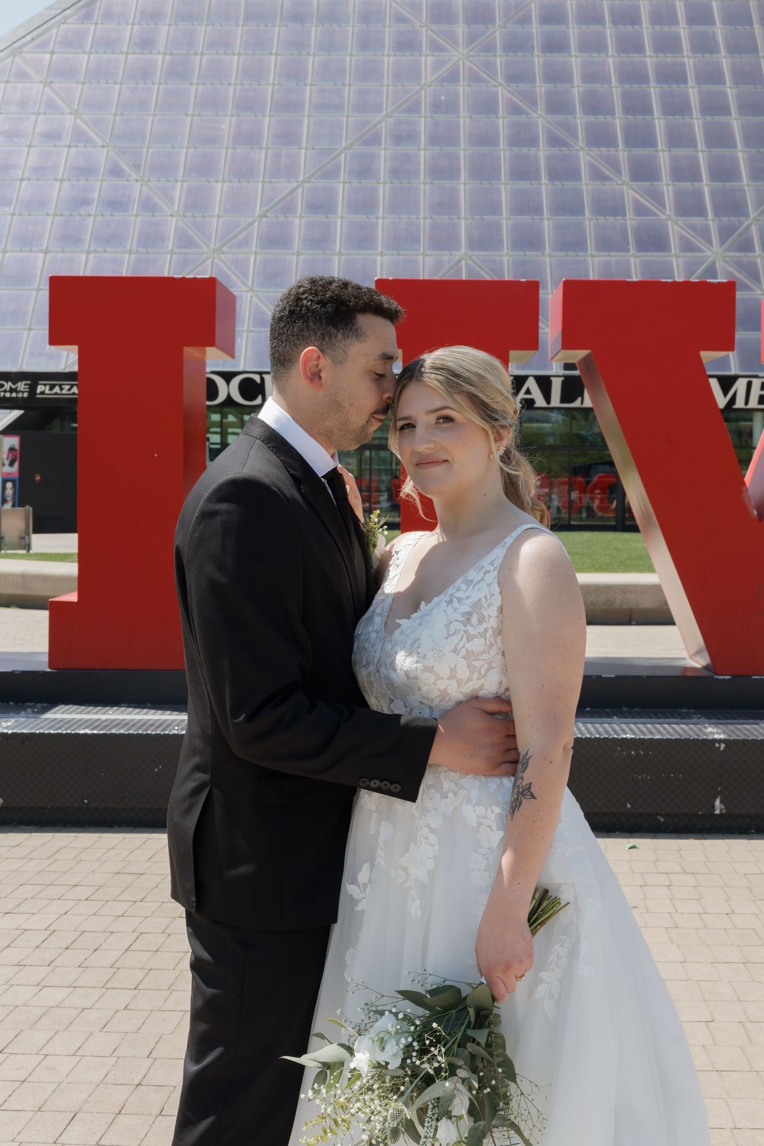 A couple in wedding attire poses in front of large red letters on a sunny day, with a glass structure in the background.