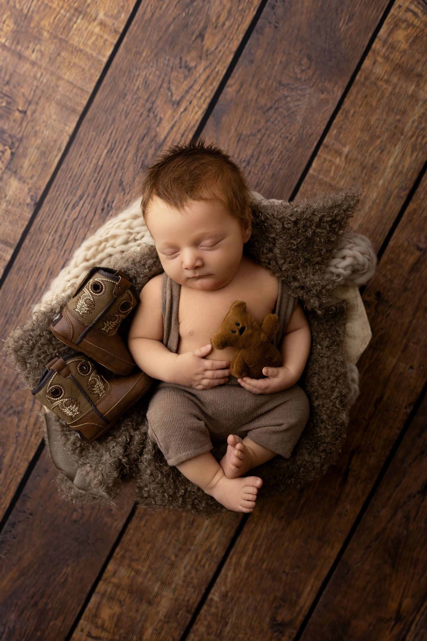 Newborn baby boy sleeping in a bowl with brown and white fabric while holding a brown bear with mini cowboy boots next to him