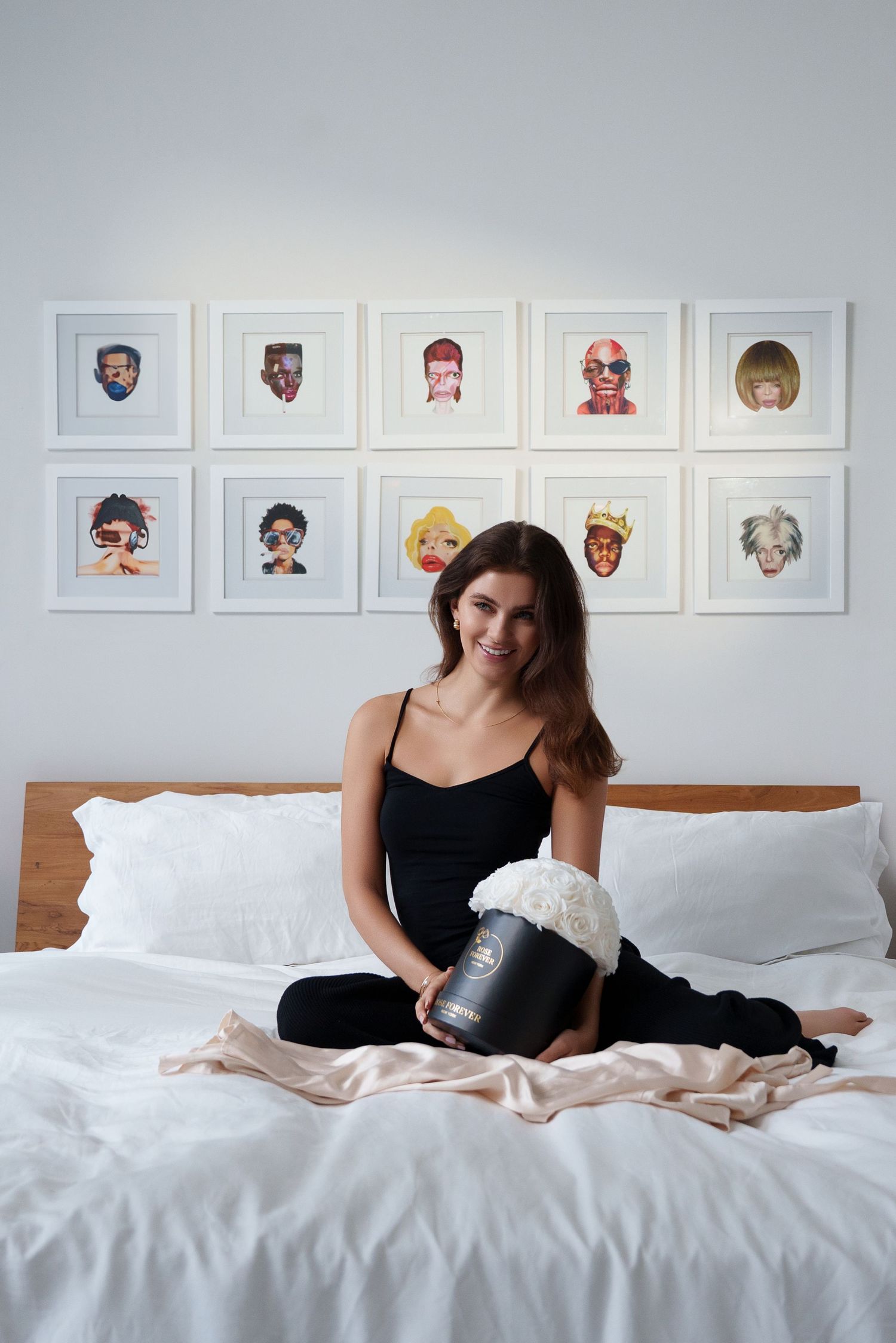 A bedroom interior with colorful portrait illustrations displayed on the wall above a white bed with neutral linens.