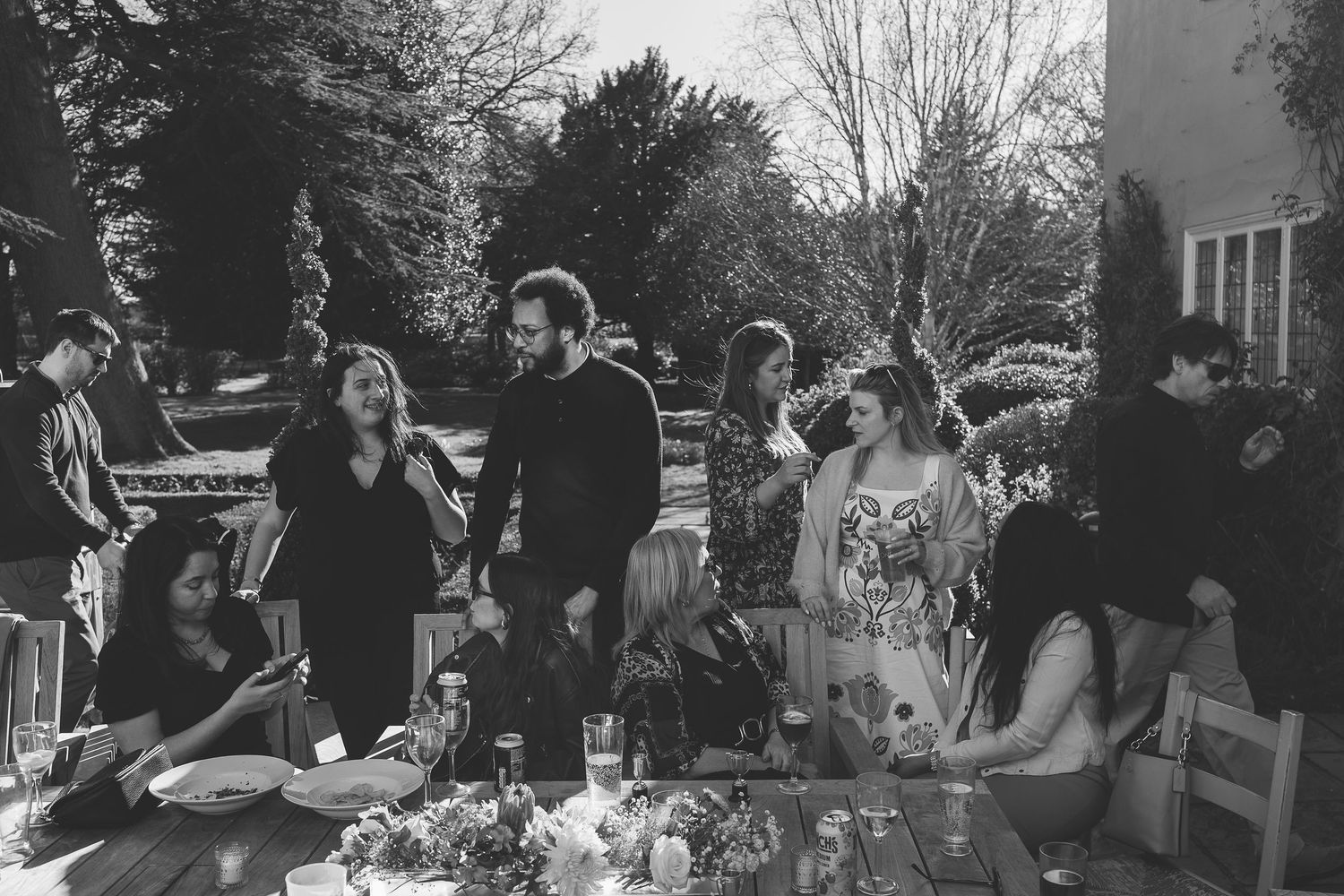 Black and white photo of people gathered outdoors at a casual garden party with refreshments on tables.