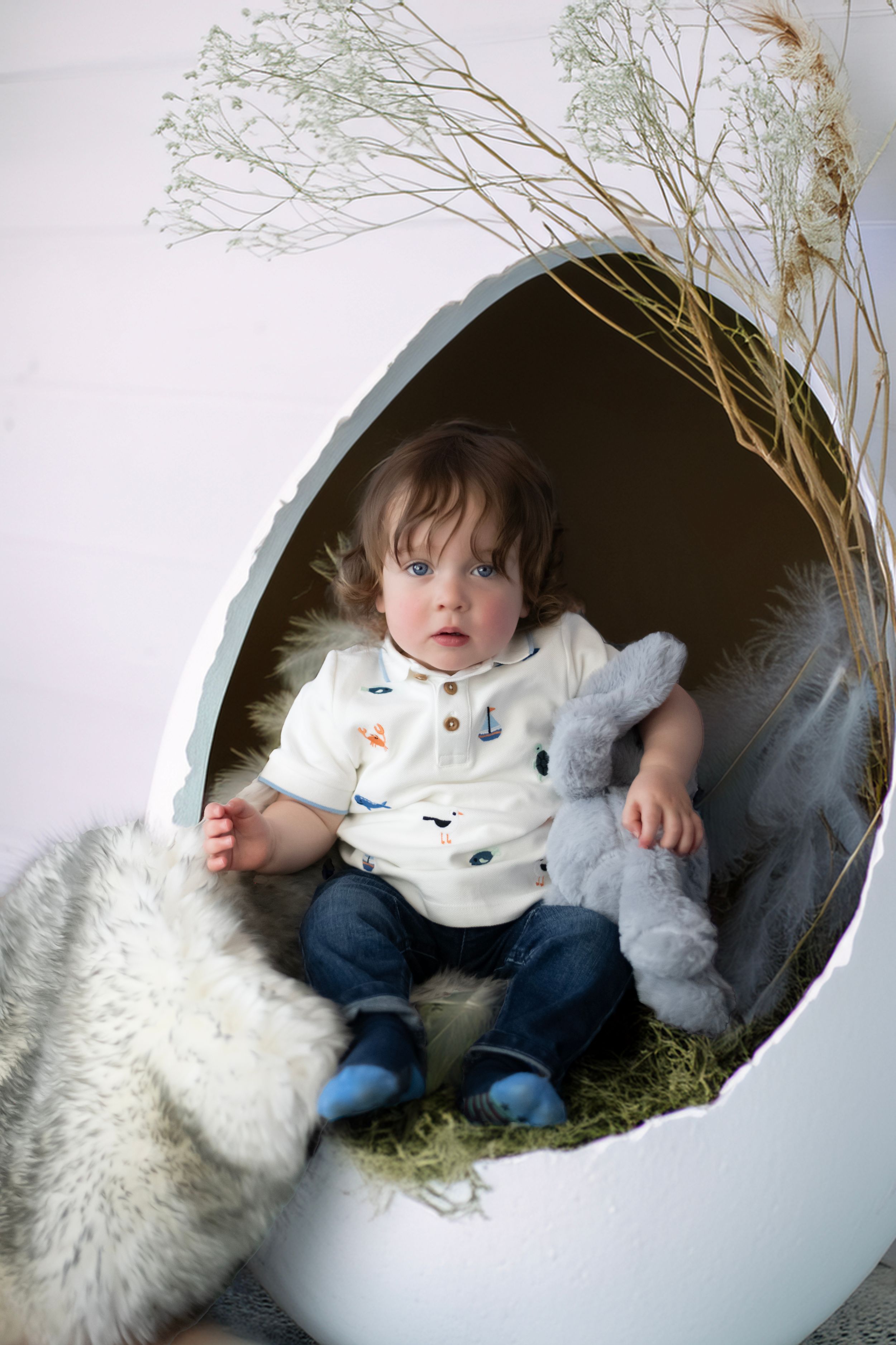 Small child relaxing in white egg-shaped chair with stuffed bunny and dried grass decorations.