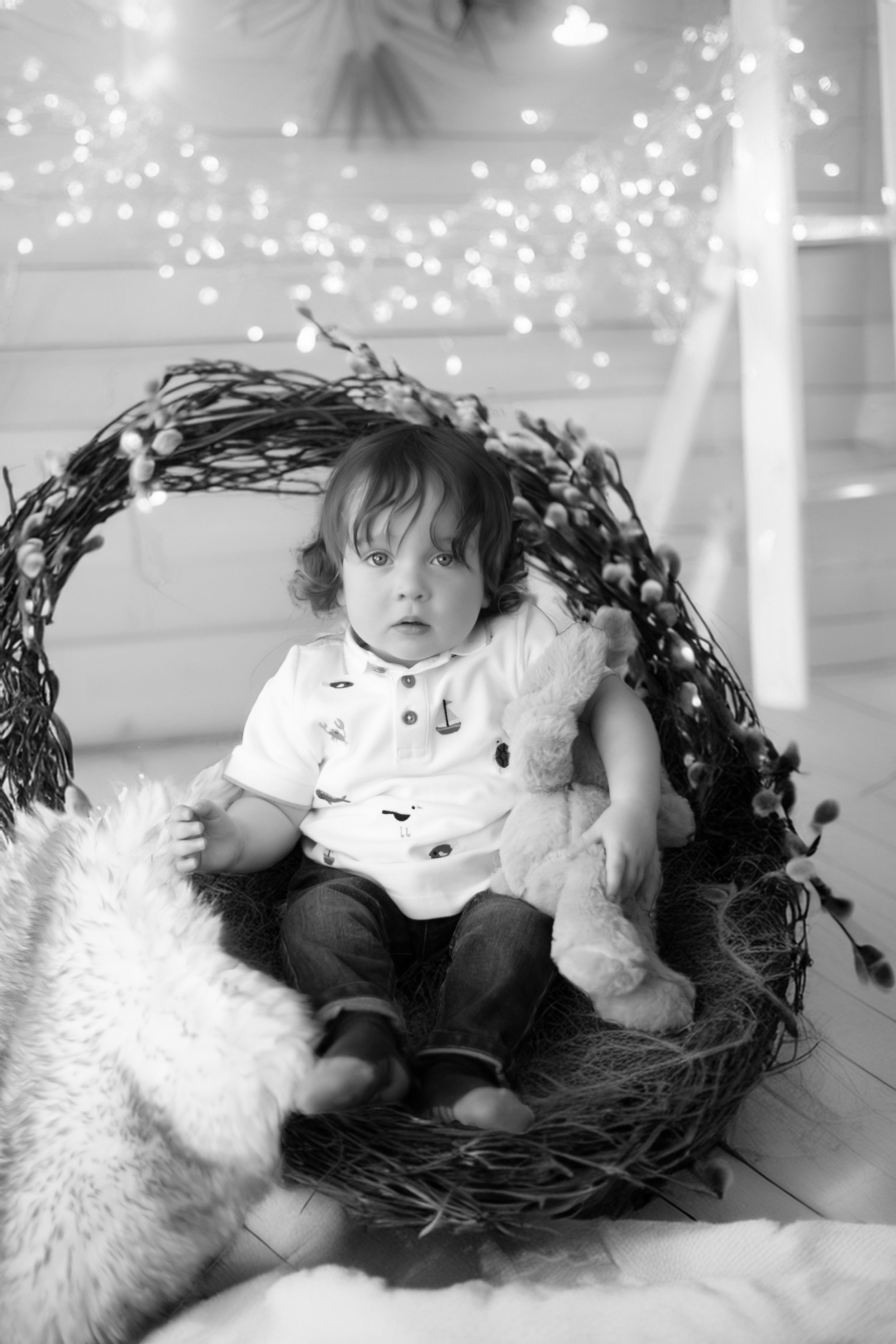Black and white photo of a toddler sitting in a twig wreath nest with a stuffed bunny and twinkle lights in background.