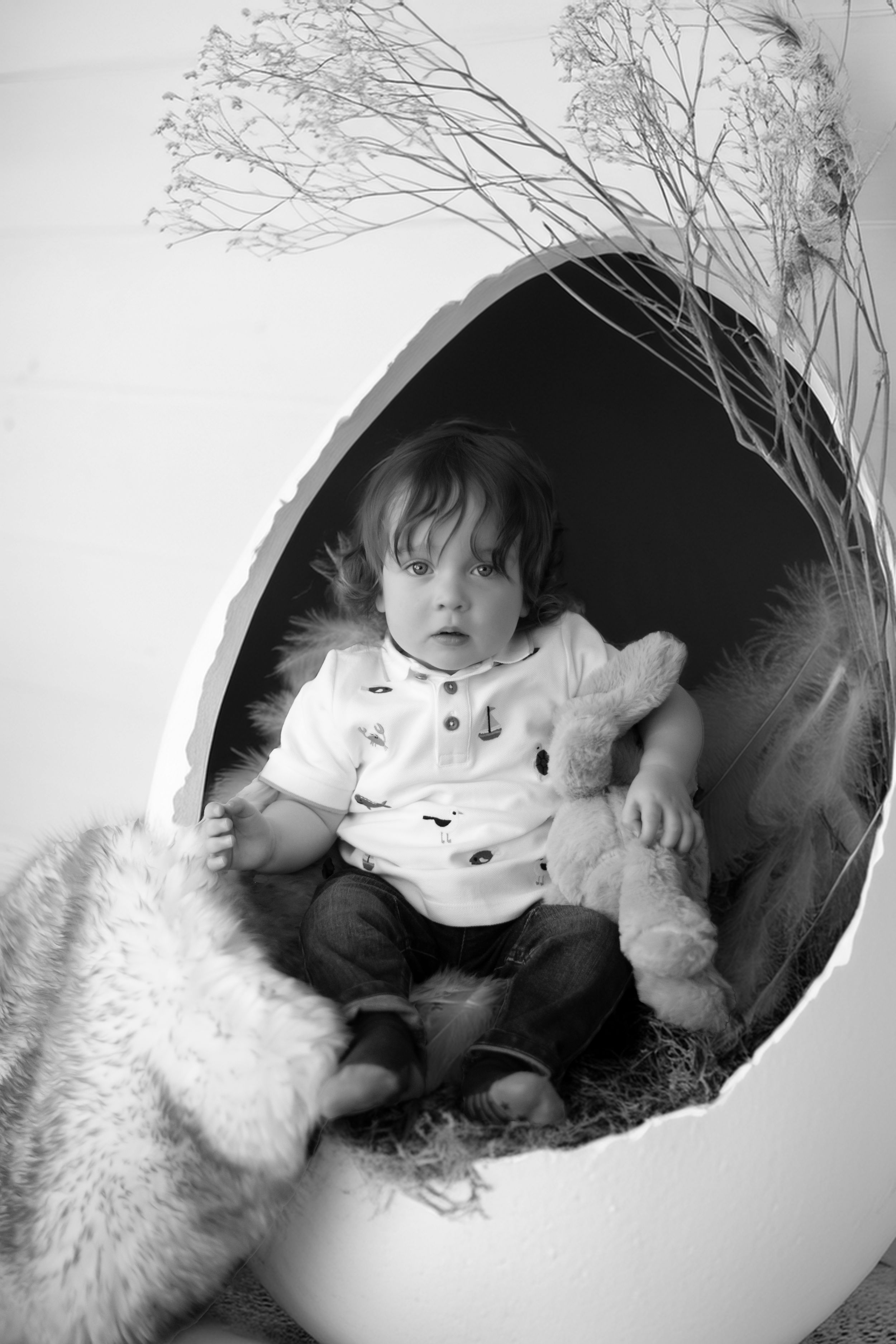 Black and white photo of a small child sitting in a white egg-shaped chair with plush toys and dried flowers.