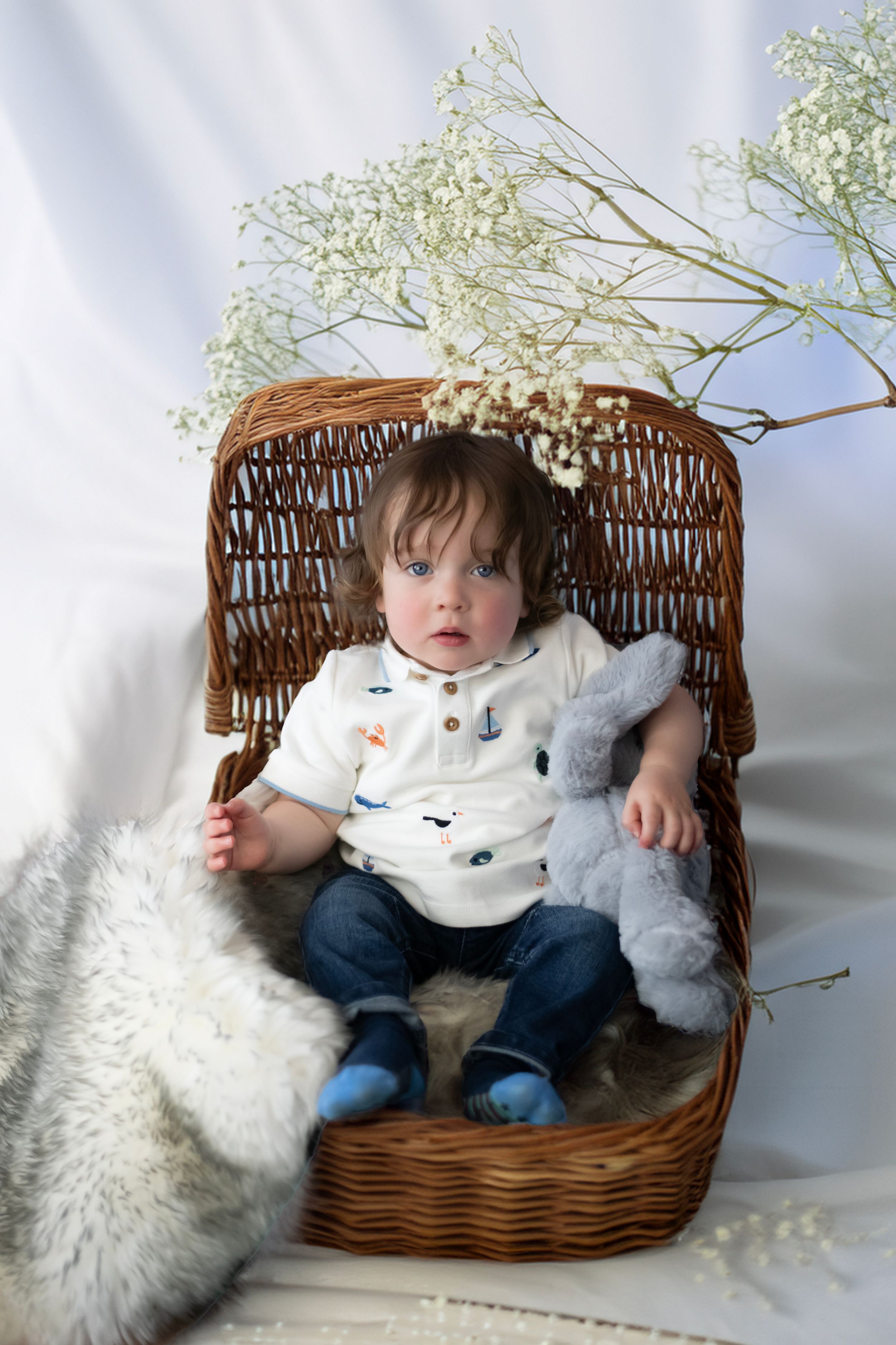 A small child sits in a woven wicker chair with a stuffed bunny toy and baby's breath flowers in a dreamy setting.