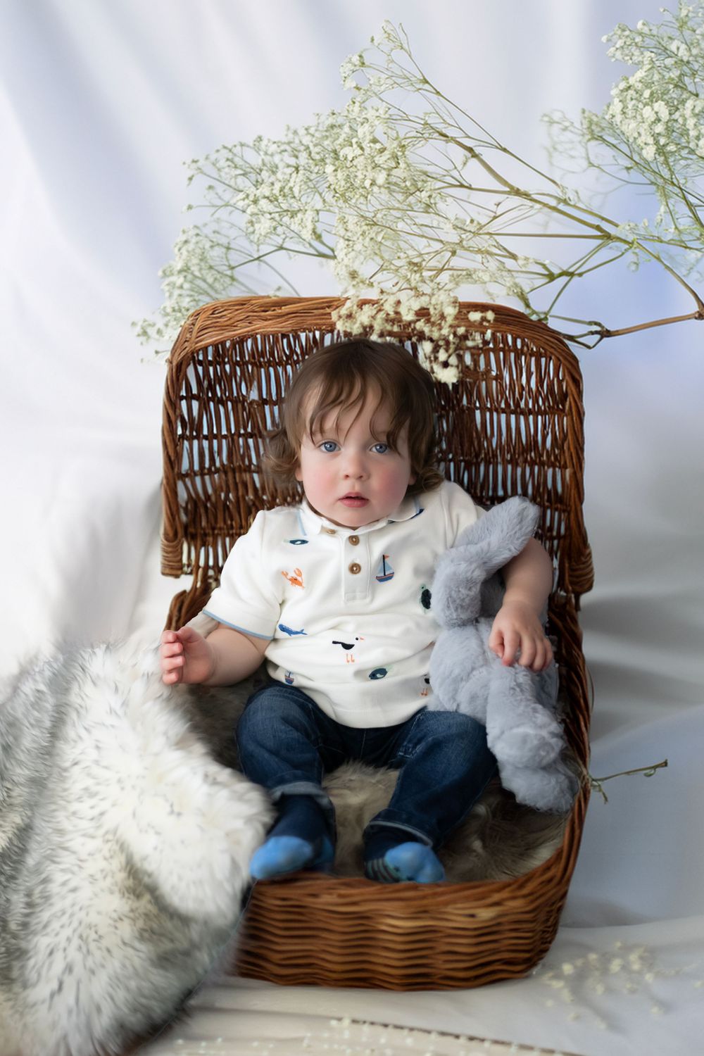 A small child sits in a woven wicker chair with a stuffed bunny toy and baby's breath flowers in a dreamy setting.