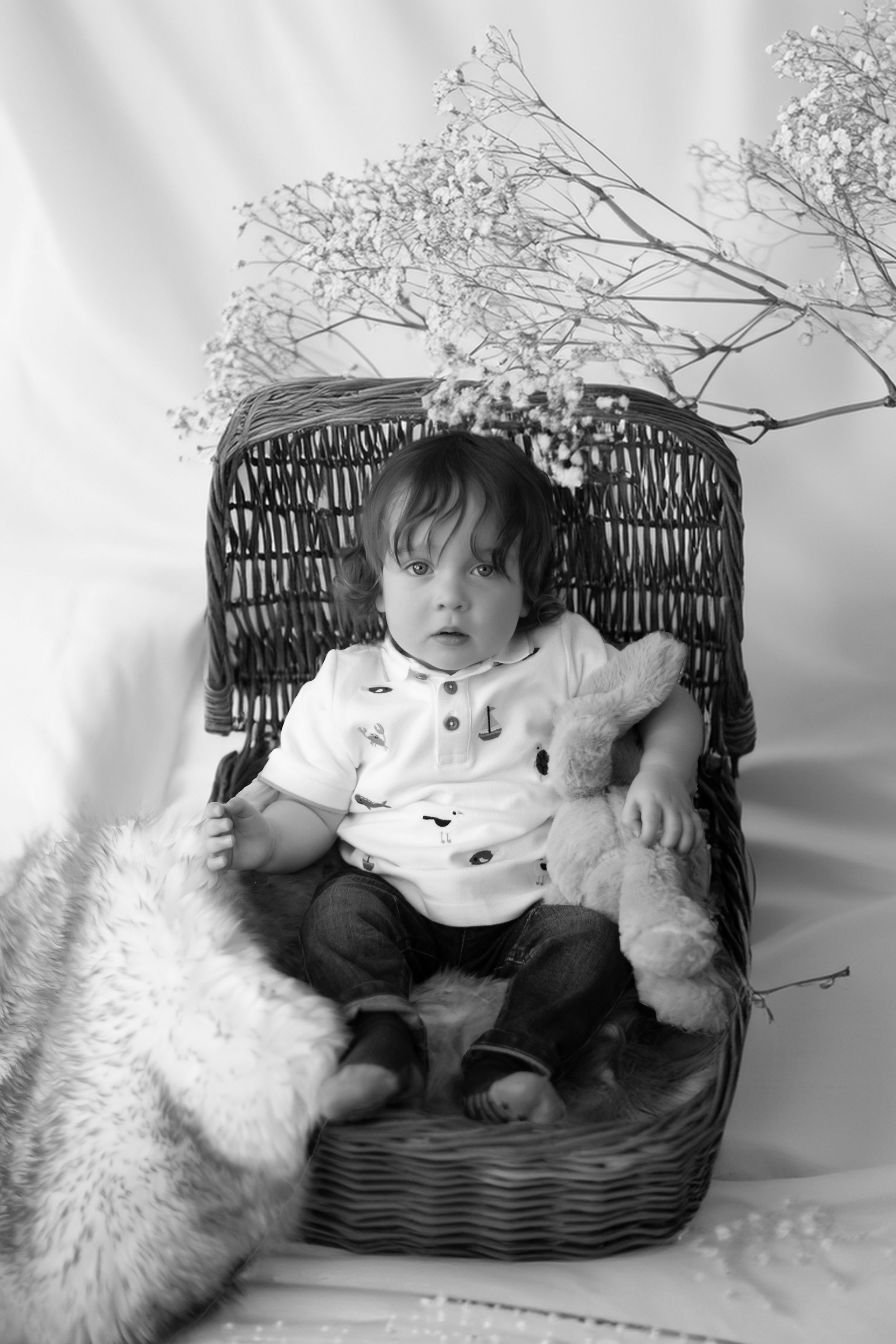 A young child sits in a wicker chair holding a stuffed bunny toy while a small pet looks on in black and white.