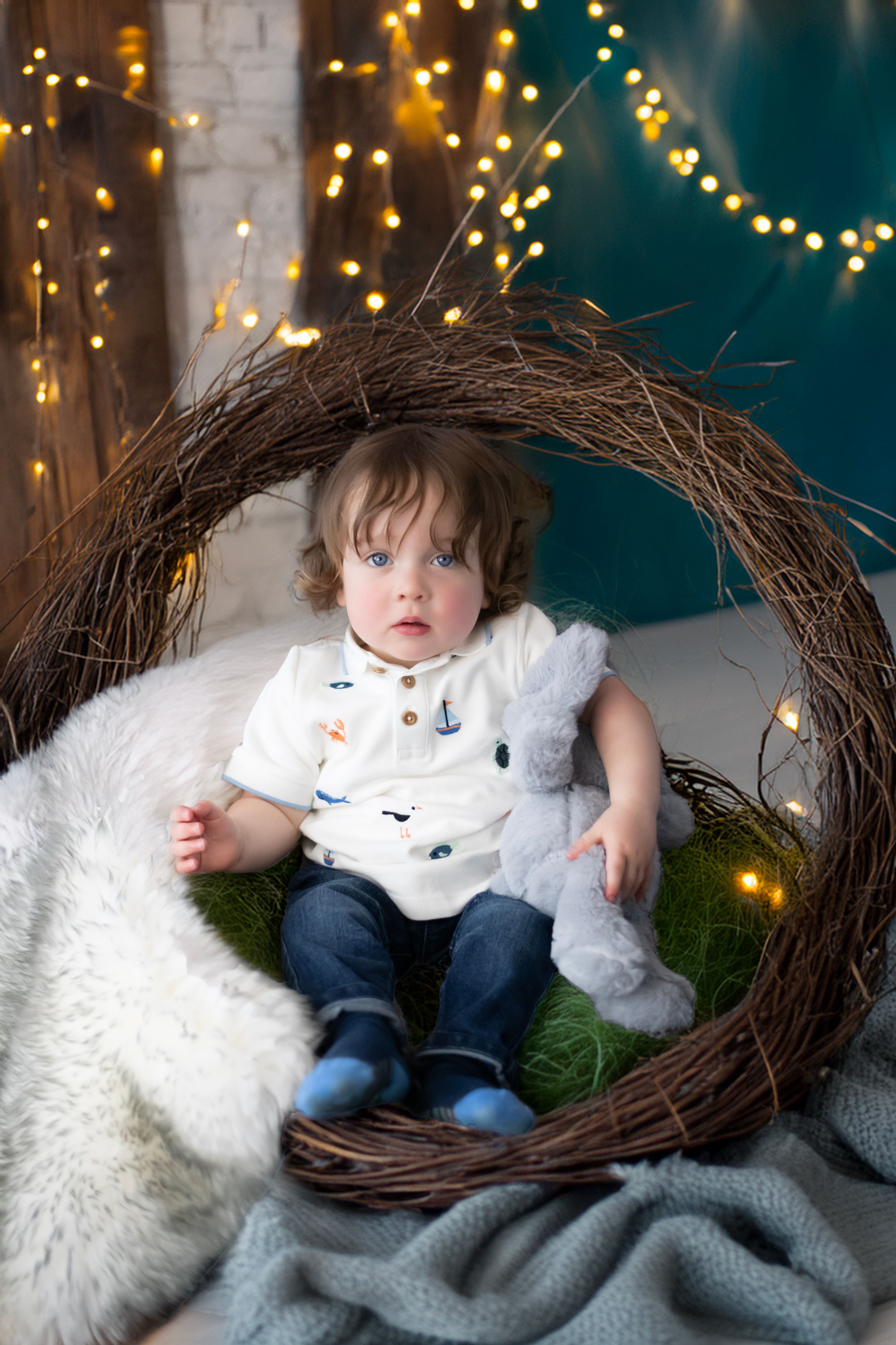 A child sits in a large twig wreath surrounded by string lights and cozy decor against a teal background.