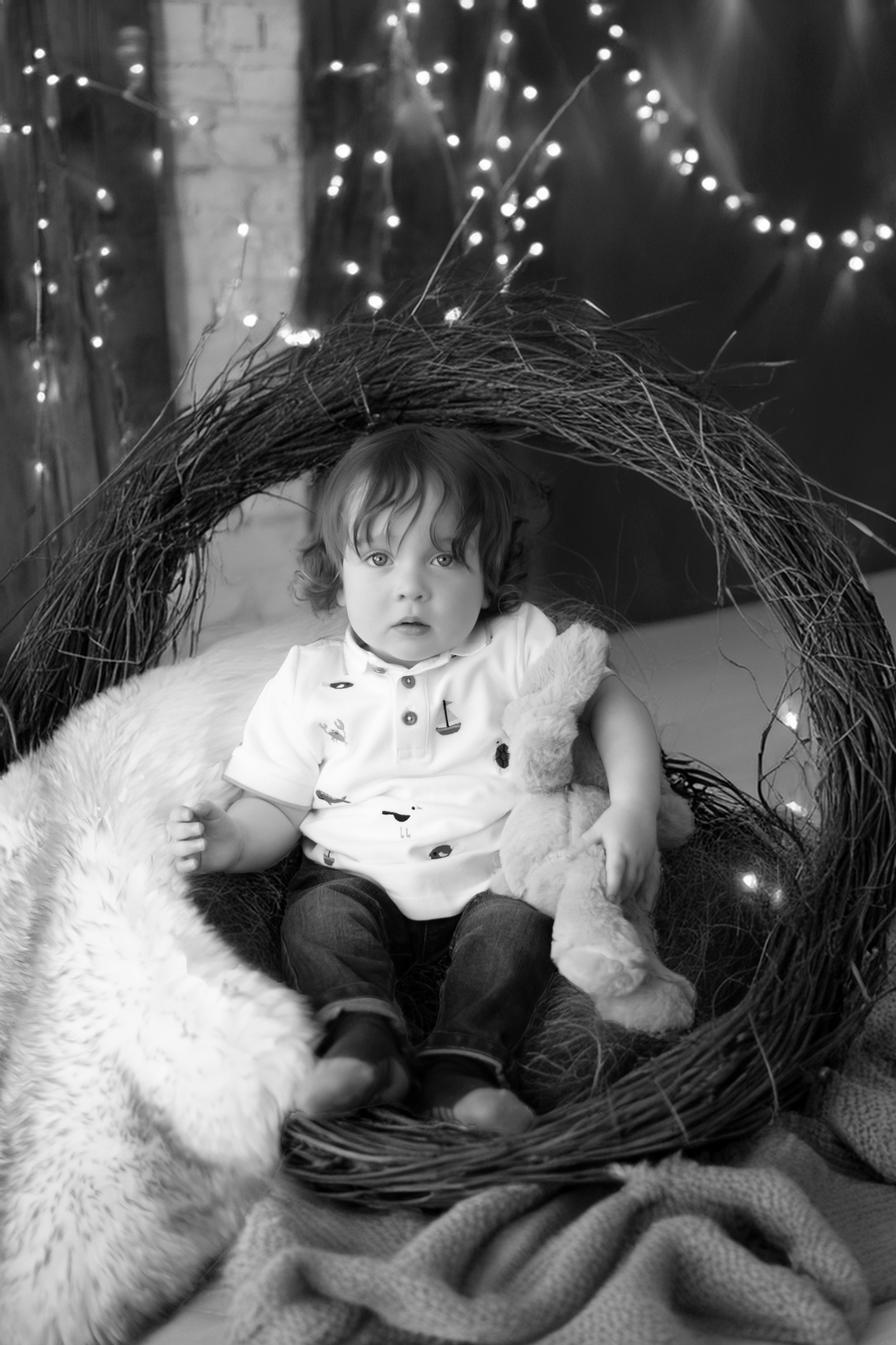 Black and white portrait of a baby nestled in a twig wreath with twinkle lights and a stuffed animal.