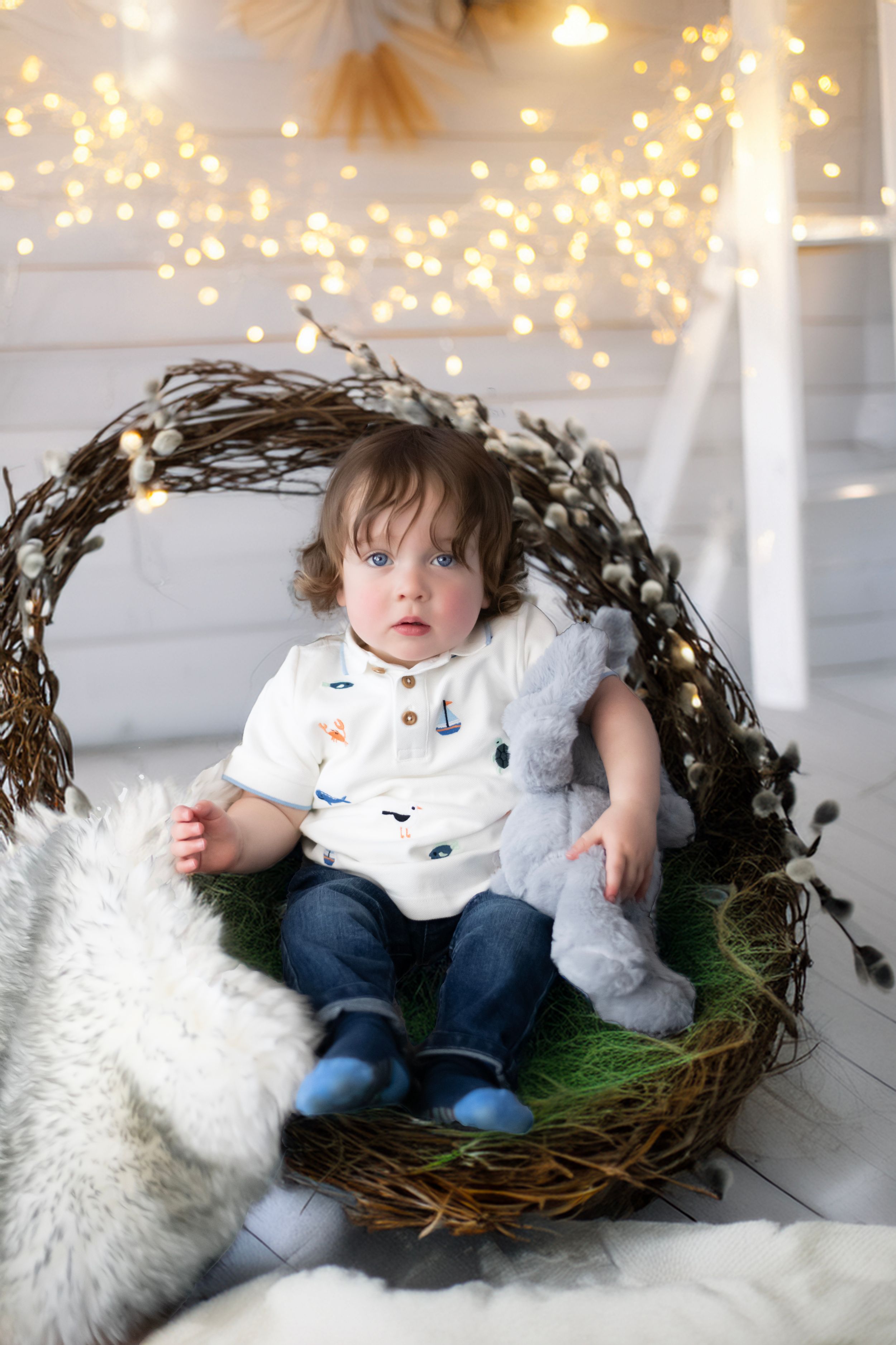 Small child sits in decorative nest basket with twinkling lights and white fur decor in dreamy holiday setting.