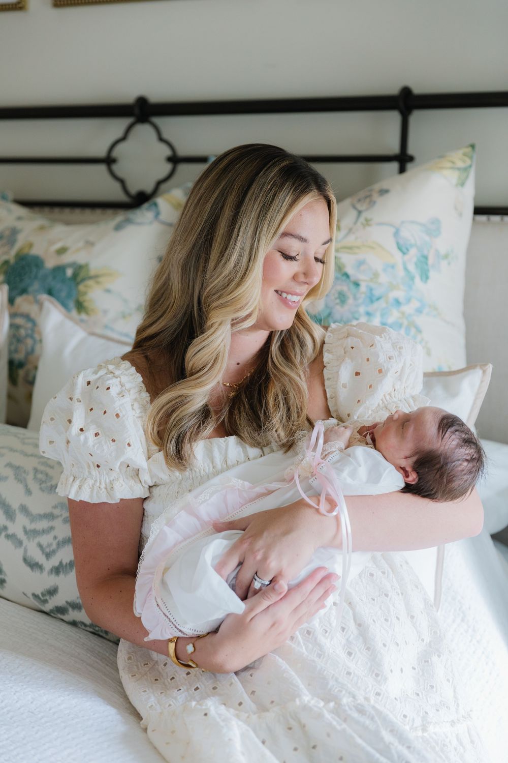 A mother in a white eyelet dress sits on a bed holding her sleeping newborn baby in white swaddle clothing.