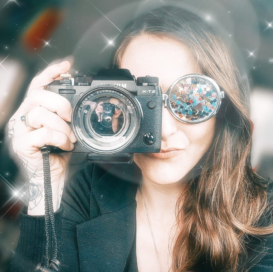 A photographer wearing round sparkly glasses holds up a vintage film camera while smiling behind the lens.