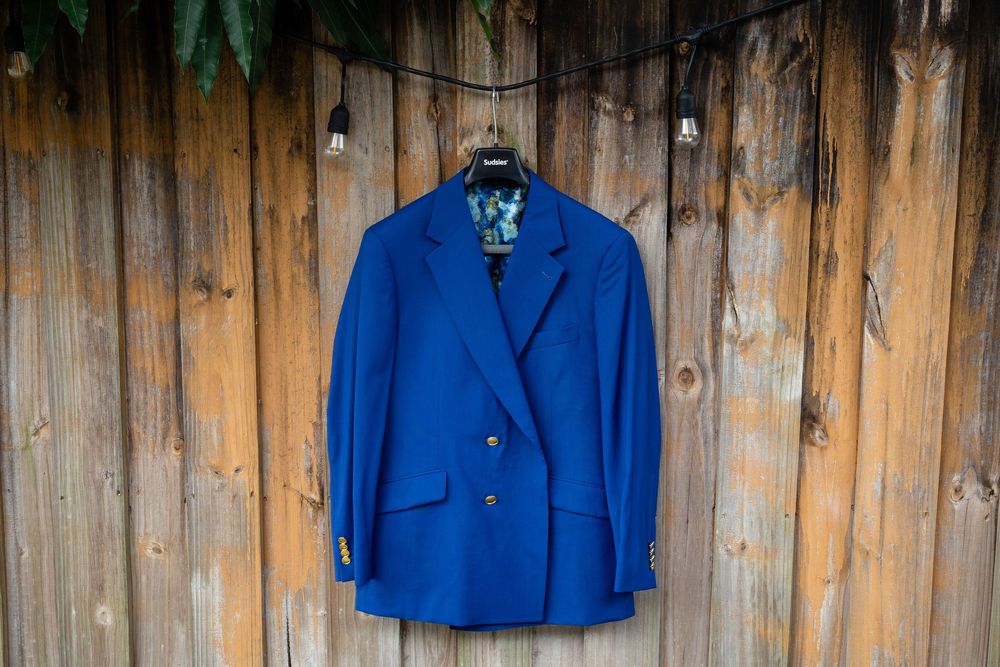 A bright royal blue double-breasted blazer hangs on a wooden fence backdrop.