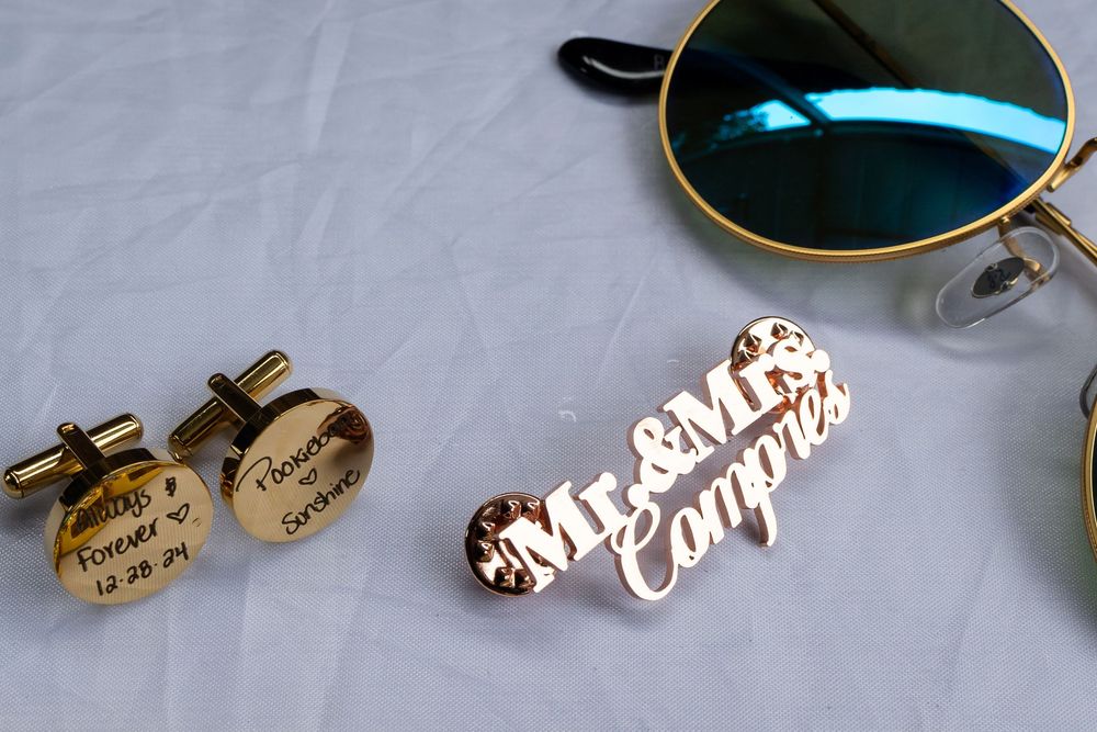 Wedding accessories including gold cufflinks and a name card displayed on white fabric next to sunglasses.