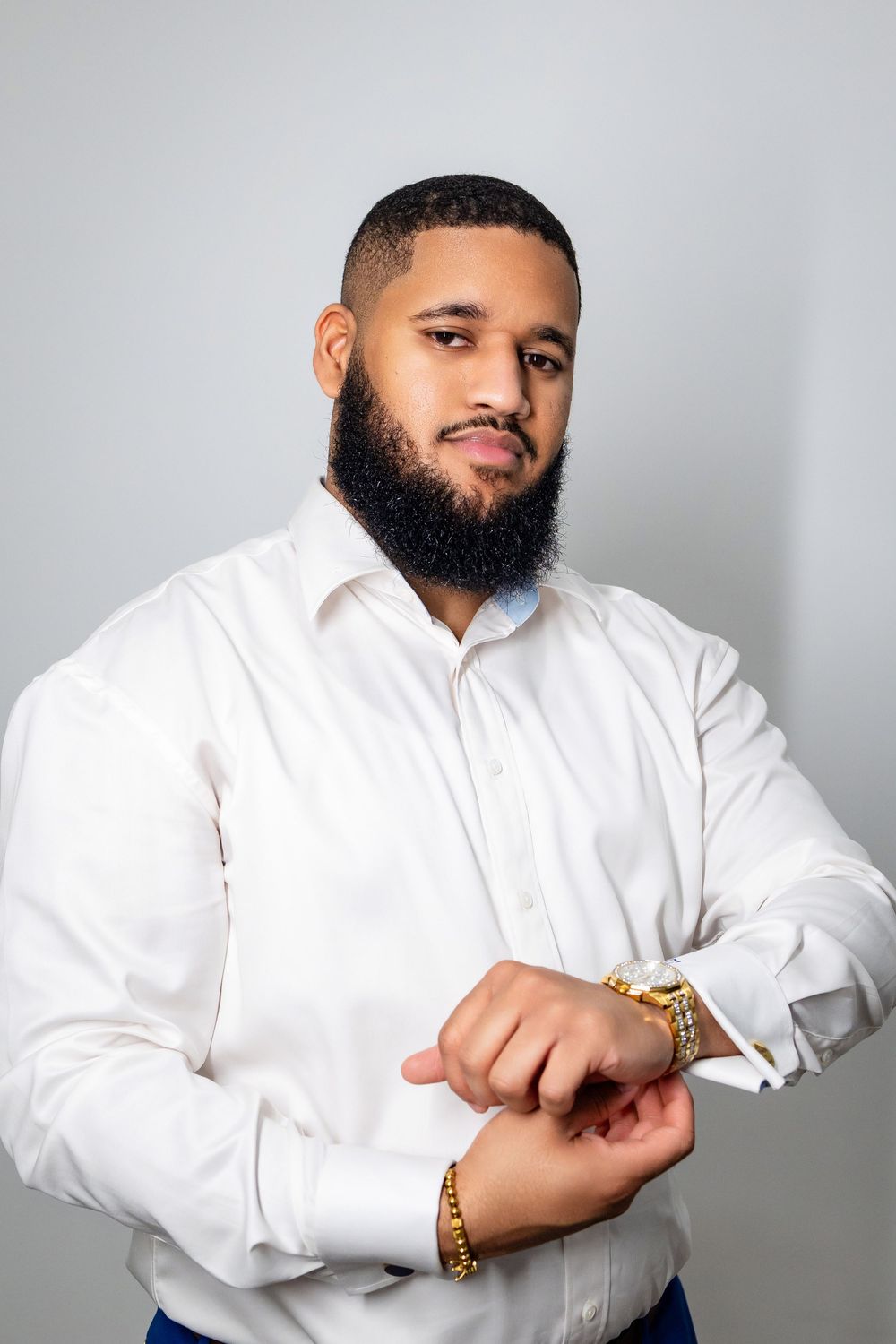 Professional studio portrait of a person in a crisp white dress shirt adjusting gold bracelets against a gray background.