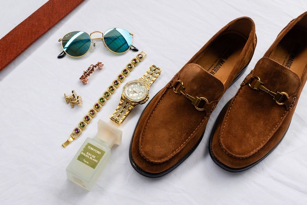 Stylish men's accessories flat lay with brown suede loafers, gold watch, sunglasses, jewelry and perfume bottle on white backdrop.