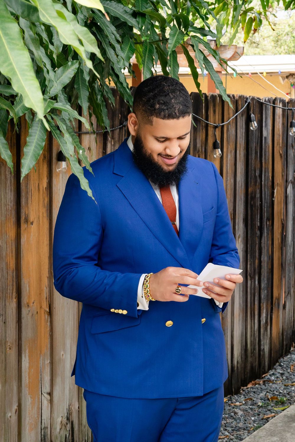 A person in a bright royal blue suit and red tie checks their phone while standing against a wooden fence.