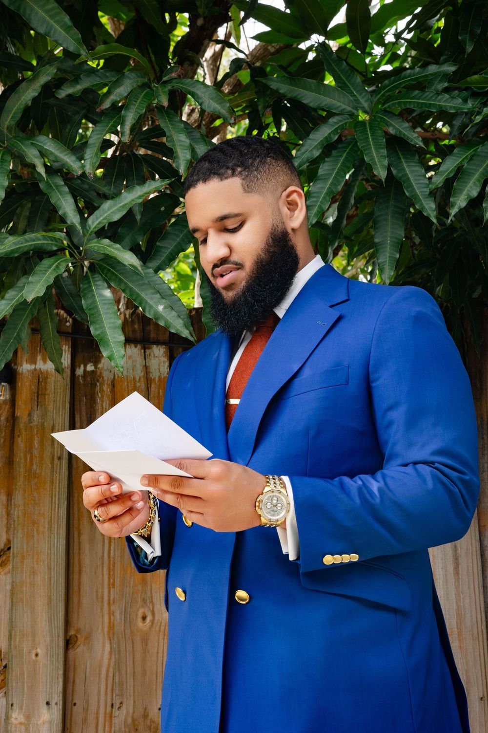 Professional in a bright blue blazer and red tie reads documents while standing outdoors near tropical foliage.