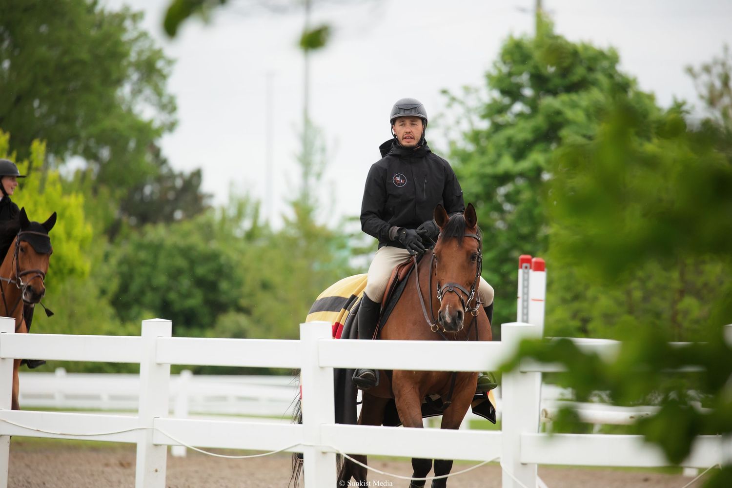 An equestrian rider in black attire jumps their chestnut horse over a white fence rail during outdoor competition.